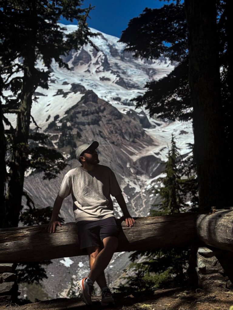 A person sitting on a wooden log in a forest with Mount Rainier and its glaciers visible in the background on a clear day.