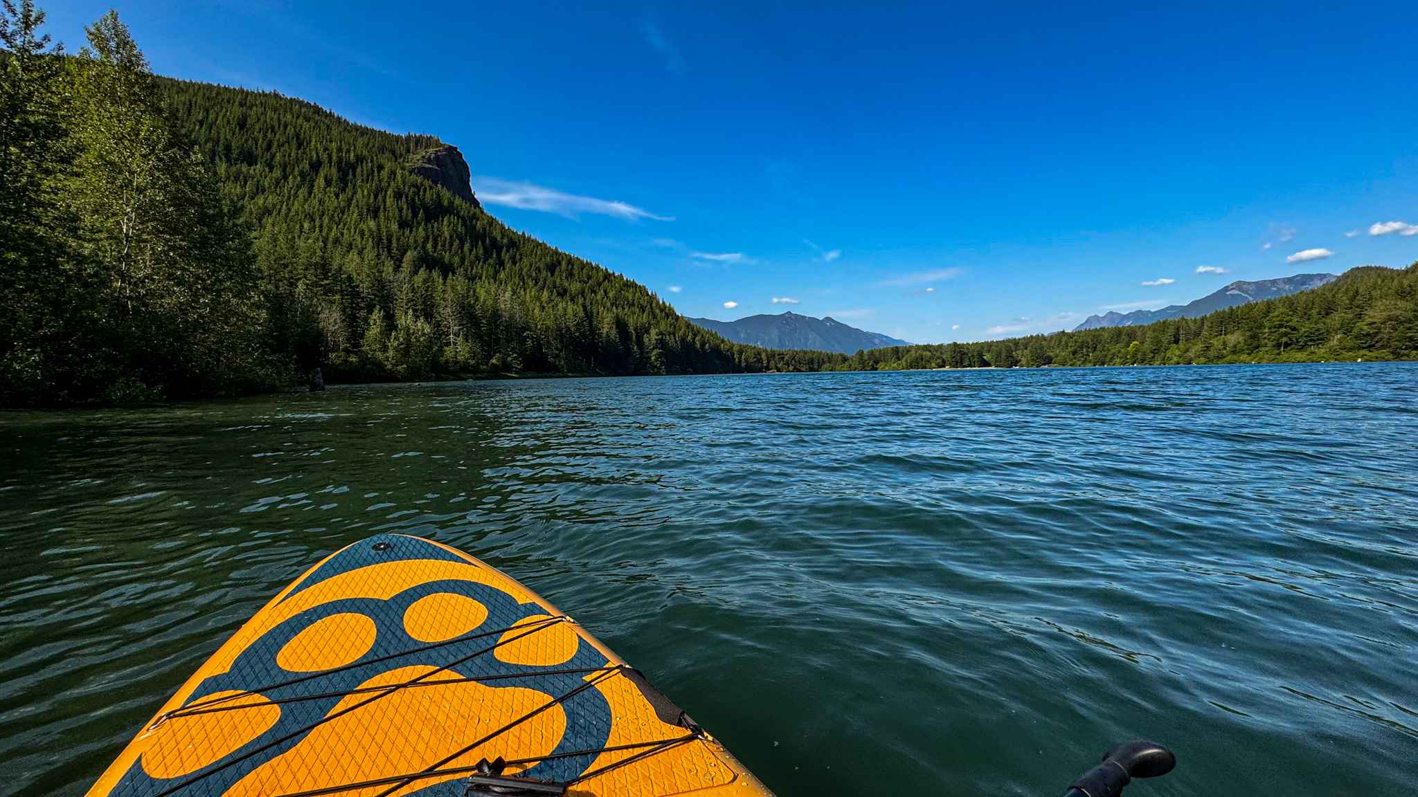 View from a paddleboard floating on a mountain lake surrounded by forested hills and distant mountains on a sunny day.