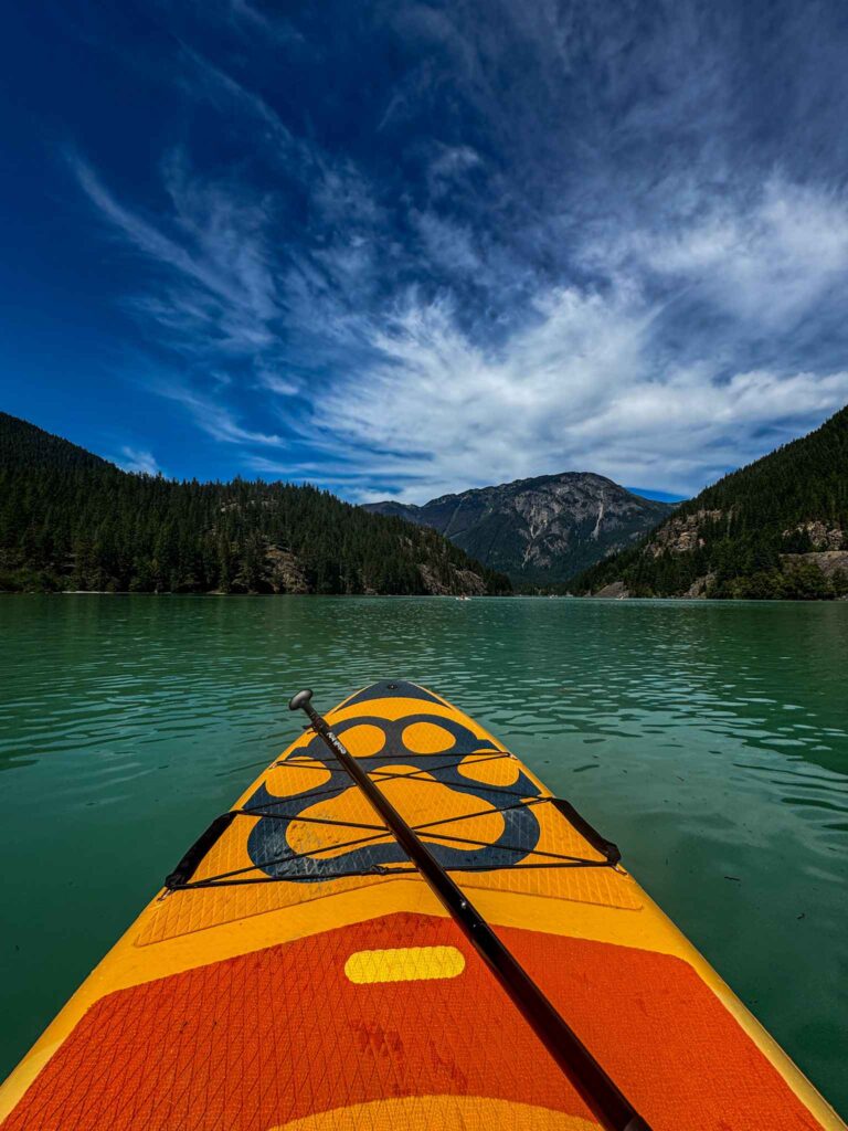 View from a paddleboard on a turquoise mountain lake surrounded by forested hills and rocky mountains under dramatic clouds.