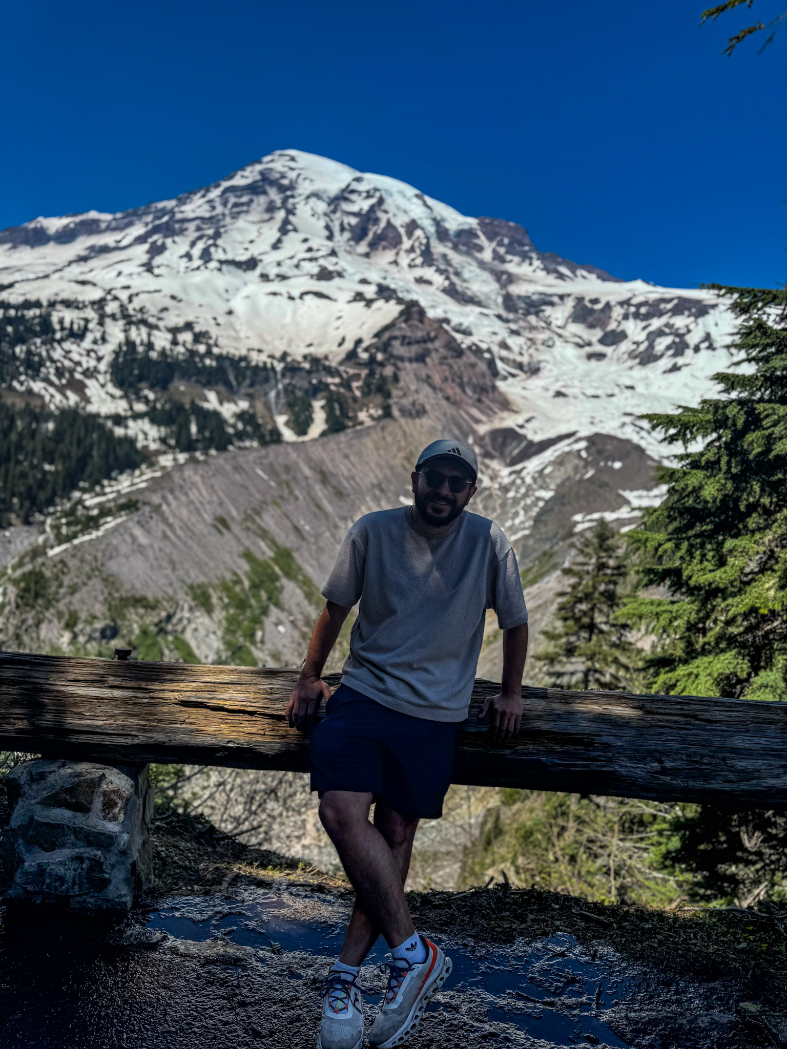 Man sitting on a wooden railing with Mount Rainier’s snow-covered peak in the background on a clear blue sky day.
