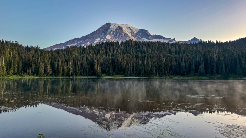 Mount Rainier reflected in a calm alpine lake surrounded by evergreen forest in Mount Rainier National Park.