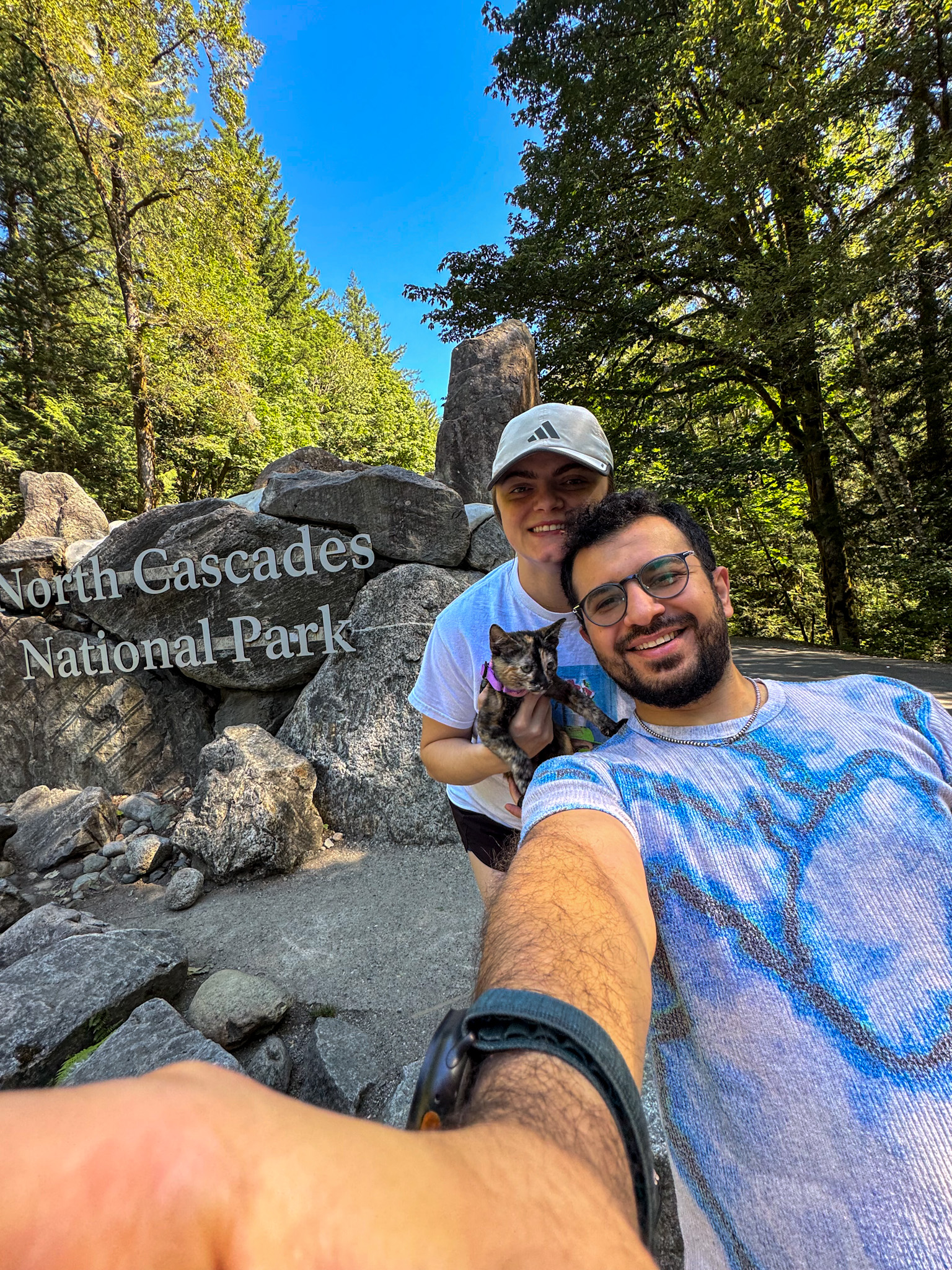Couple holding a tortoiseshell cat in front of the North Cascades National Park entrance sign in Washington.