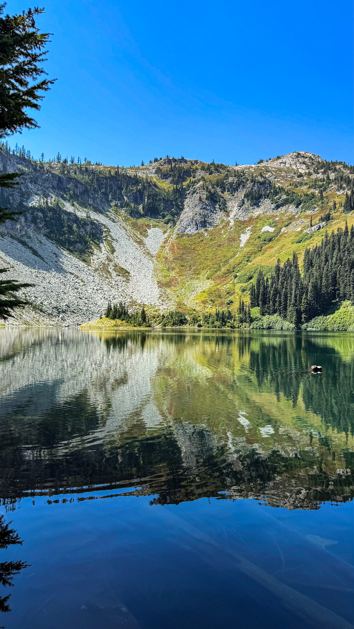 Clear alpine lake reflecting rocky mountain slopes and evergreen trees under a blue sky in Washington State.
