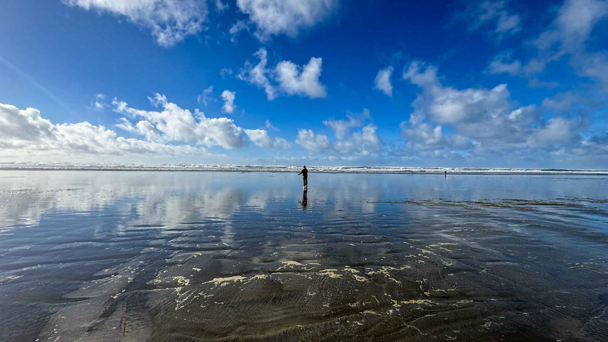 Person standing on a wide Washington beach with reflections of clouds and blue sky on wet sand at low tide.
