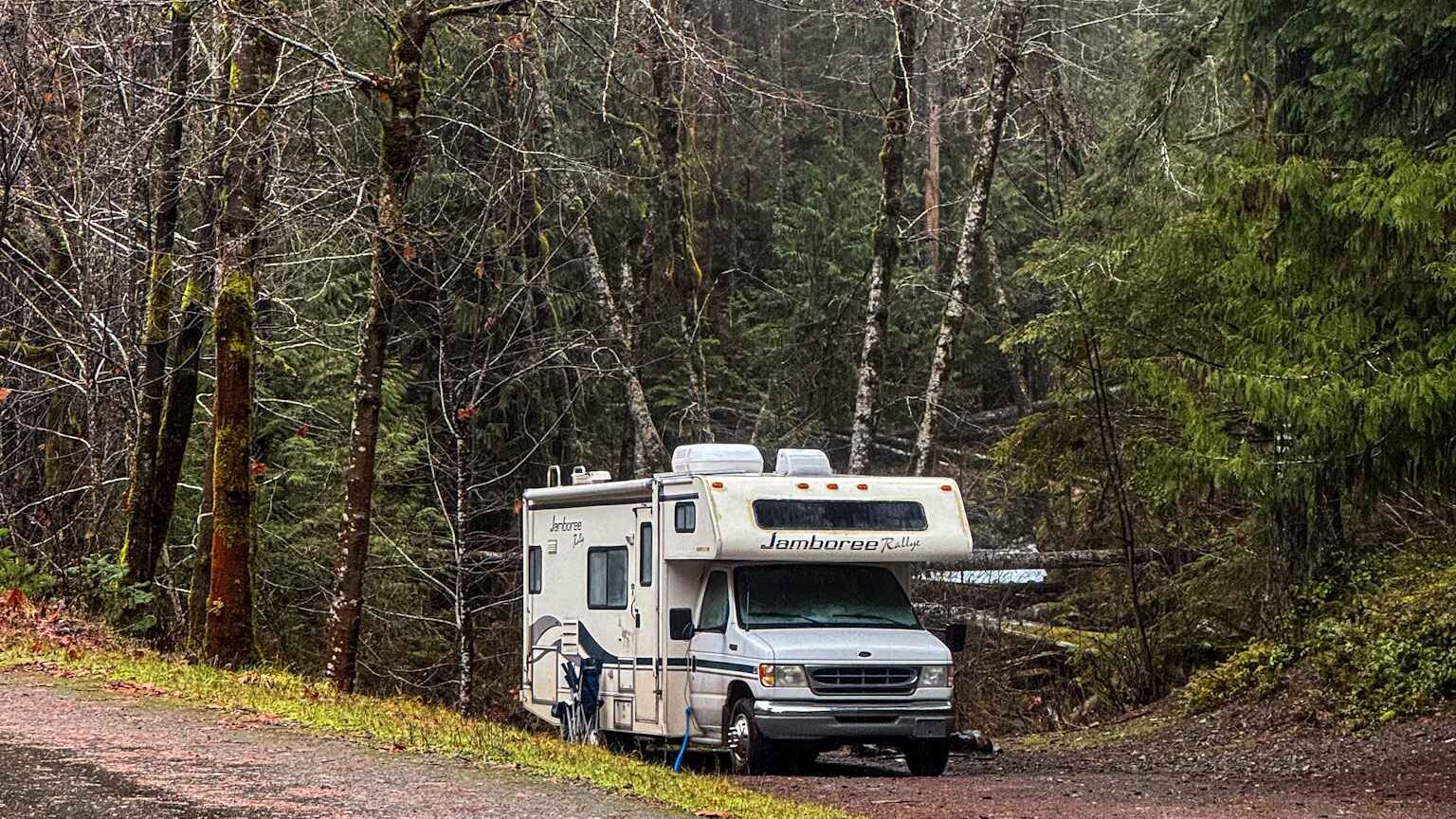 A Class C RV parked at a forest campsite surrounded by tall trees on a rainy, overcast day.