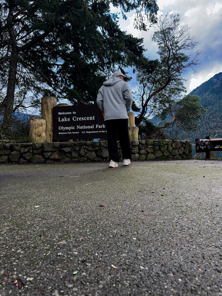 Person standing near the Lake Crescent welcome sign in Olympic National Park with forested hills in the background.
