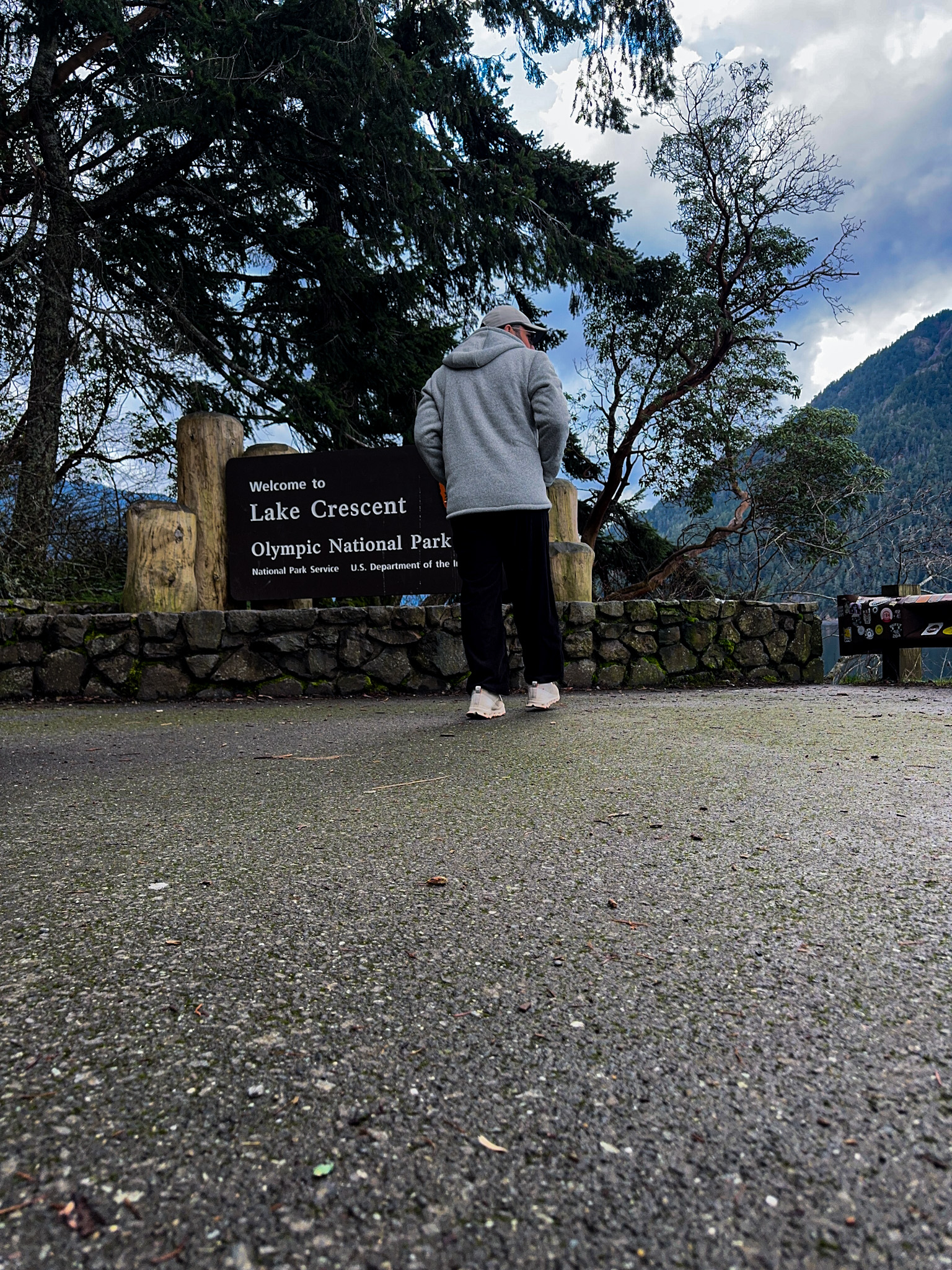 Person standing near the Lake Crescent welcome sign in Olympic National Park with forested hills in the background.
