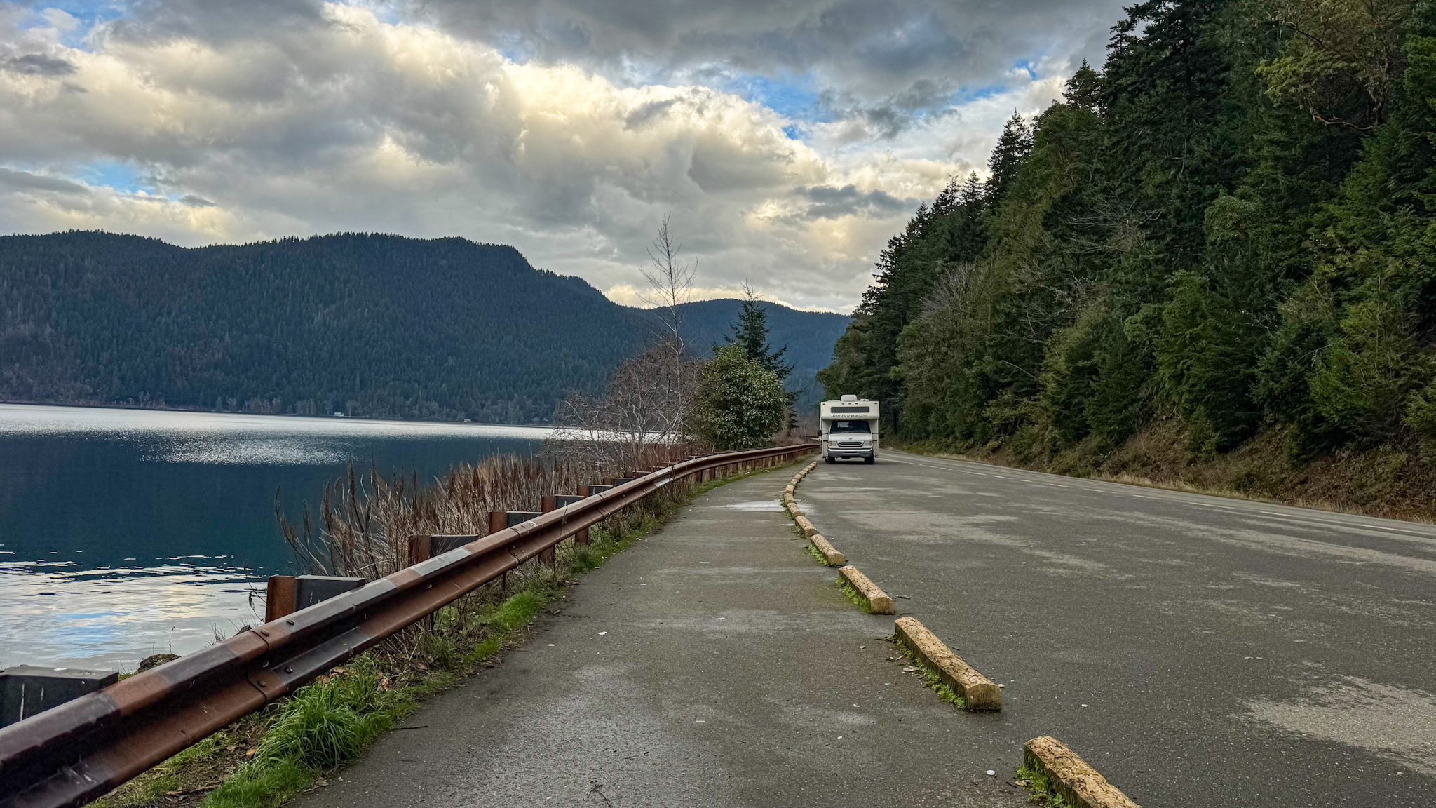 RV driving along the shoreline road next to Lake Crescent with forested hills and cloudy skies in Olympic National Park.
