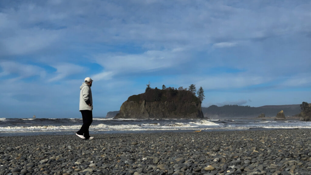 Person walking along a rocky beach with waves and a forested sea stack along the Olympic National Park coast.