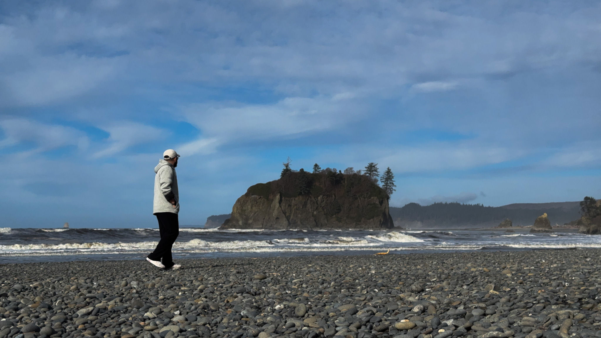 Person walking along a rocky beach with waves and a forested sea stack along the Olympic National Park coast.
