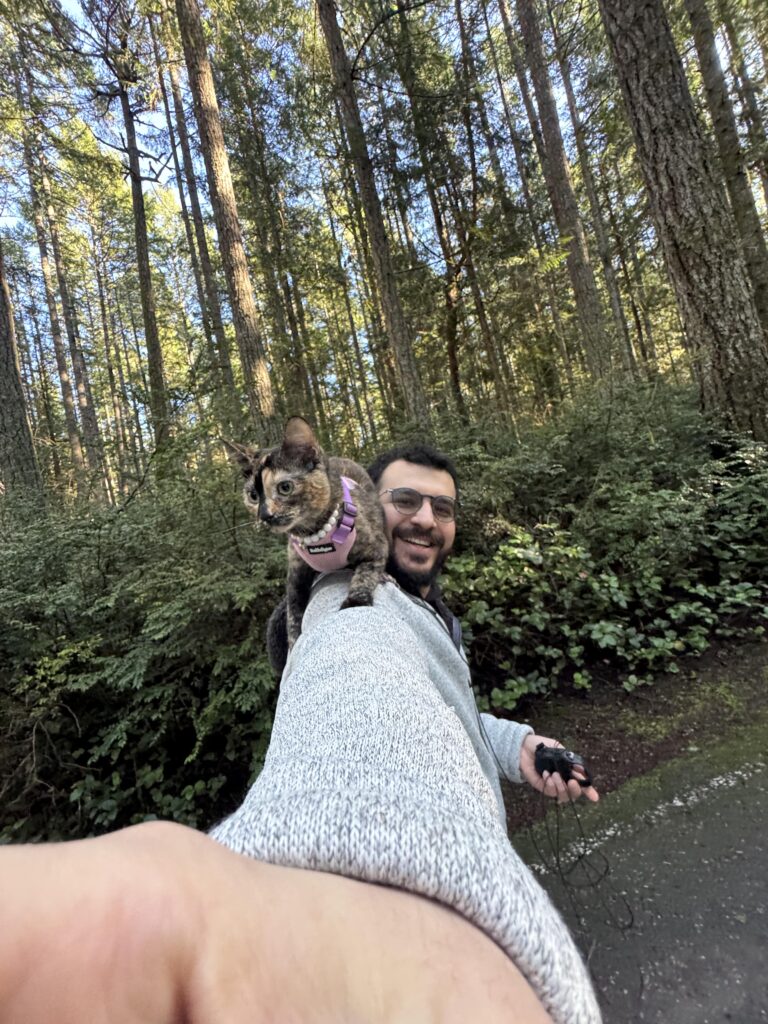 Man smiling on a forest trail in Washington with a tortoiseshell cat wearing a pink harness sitting on his shoulder.