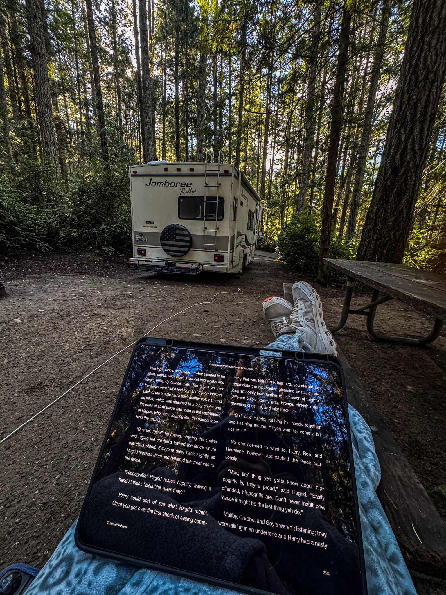 Person relaxing at a forest campsite with a tablet in the foreground and a Class C RV parked among tall evergreen trees.
