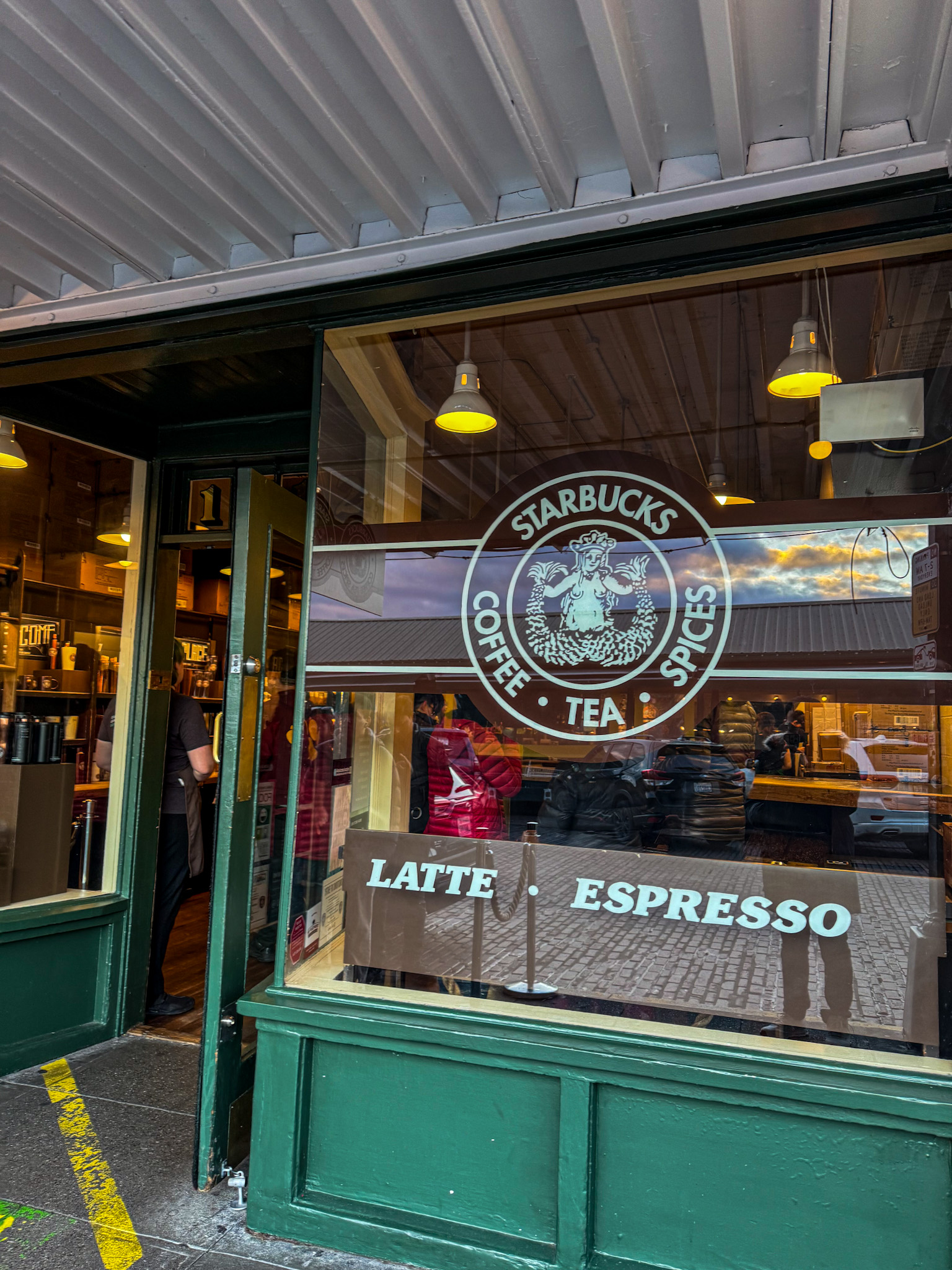 Exterior of the original Starbucks store at Pike Place Market in Seattle with the classic Starbucks Coffee Tea Spices logo on the window.
