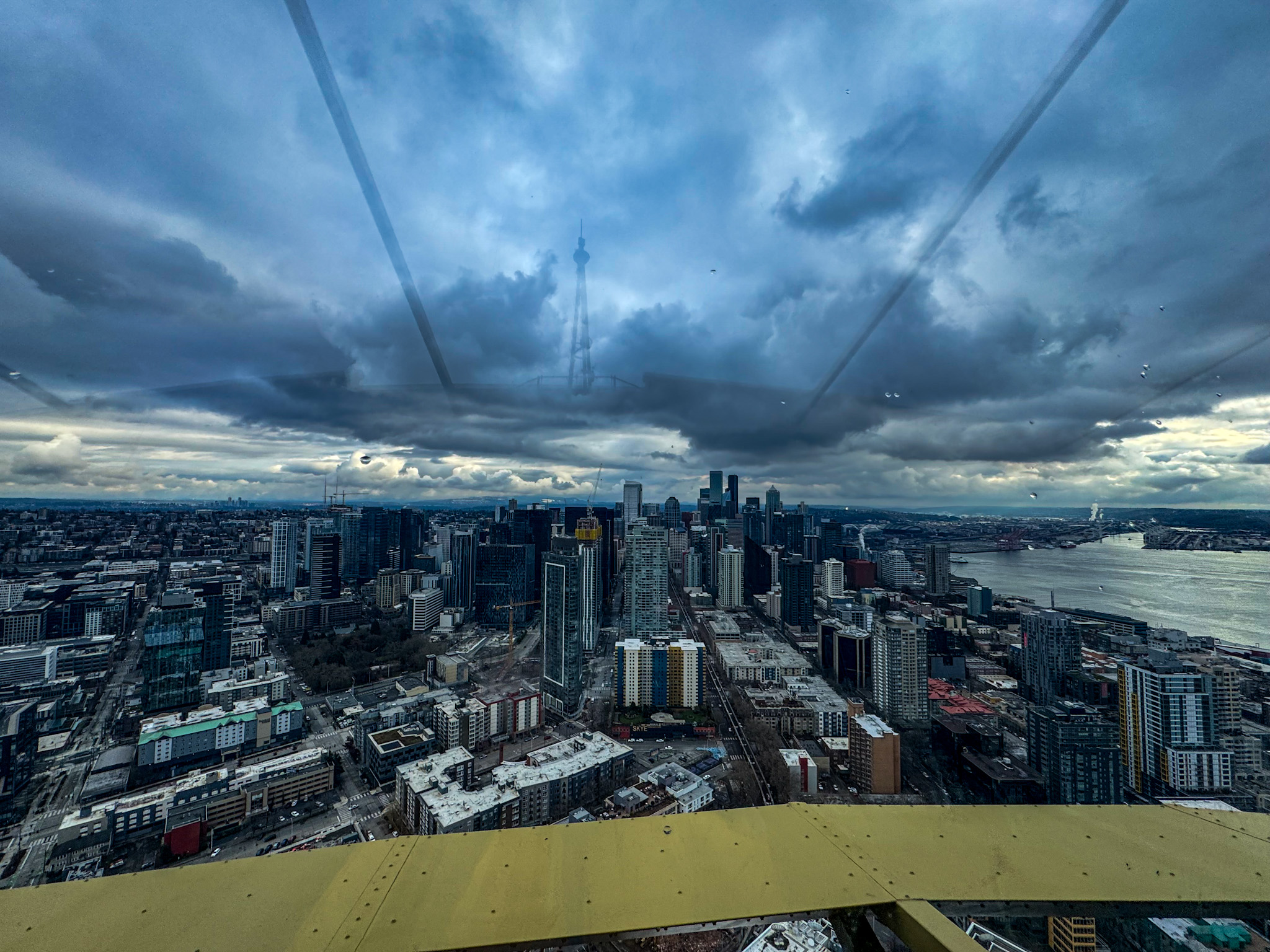 Panoramic view of Seattle skyline and Elliott Bay from the Space Needle observation deck on a cloudy day.
