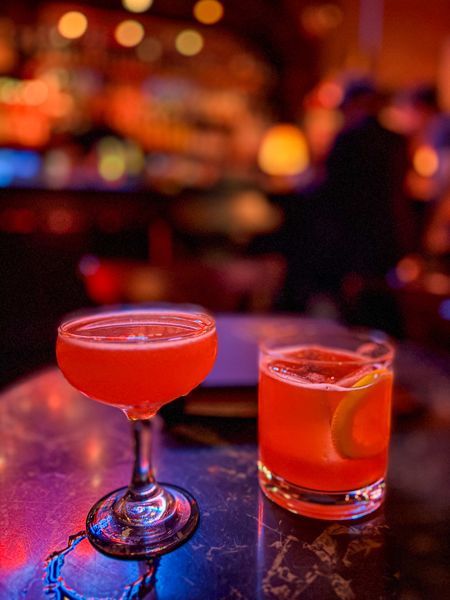 Two bright red craft cocktails on a bar table with colorful bokeh lights in the background.
