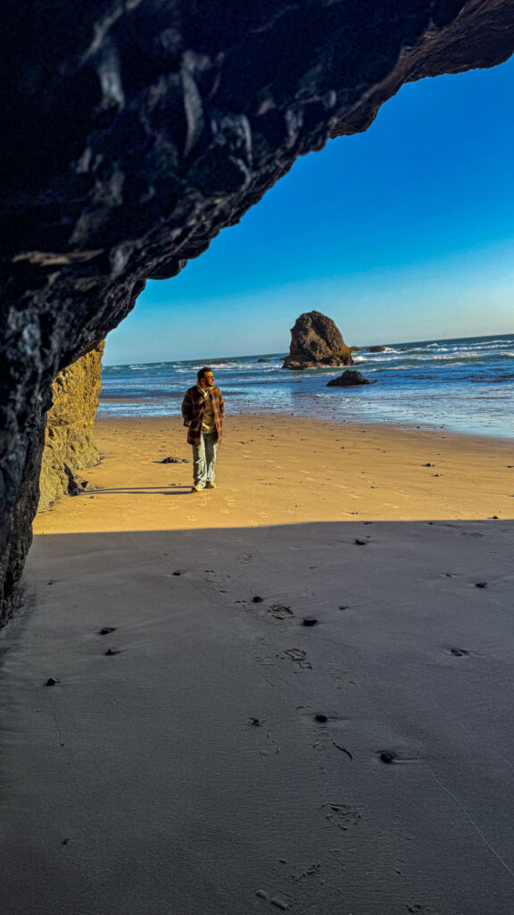 Traveler walking on a sandy Oregon Coast beach with large sea stacks and ocean waves in the background.