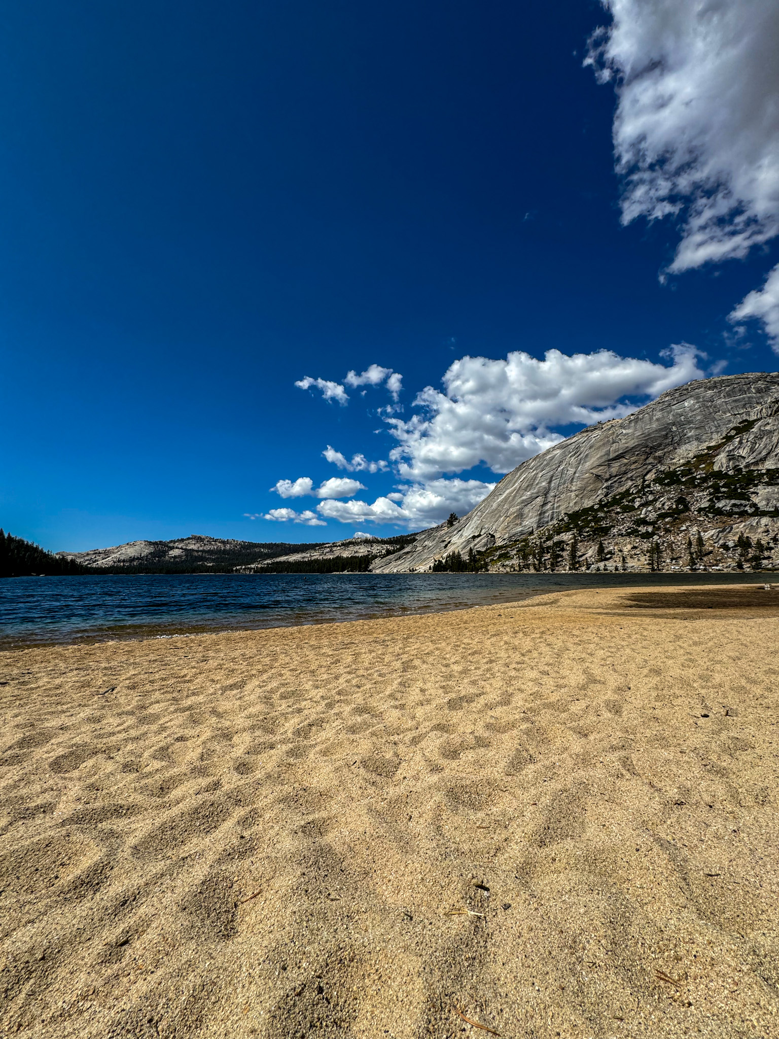 Sandy lakeshore in Yosemite National Park with clear blue water and granite dome under bright sky.