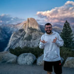 Person standing at Yosemite viewpoint with Half Dome in the background during sunset.