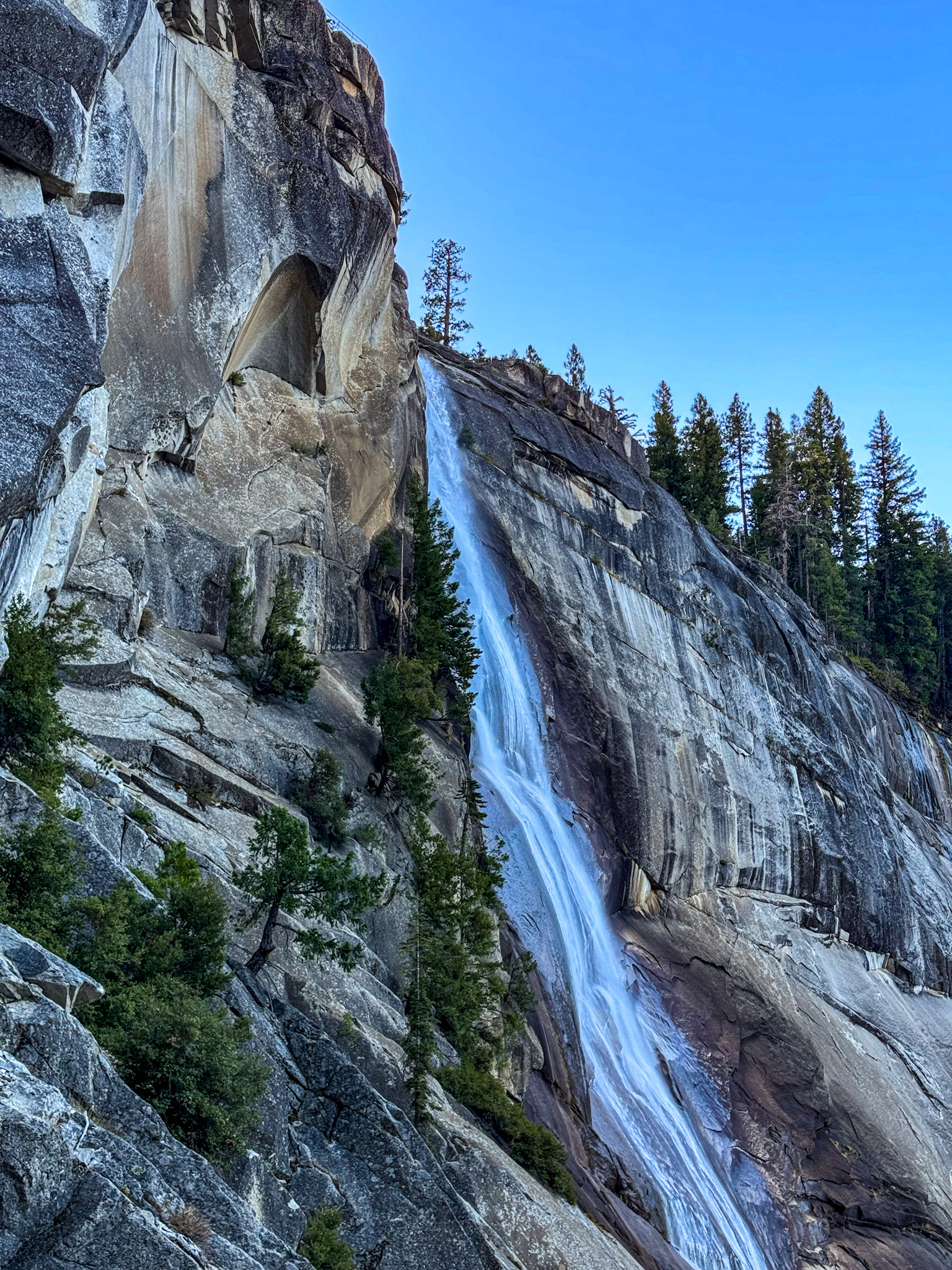 Waterfall cascading down steep granite cliff in Yosemite National Park with pine trees and clear blue sky.