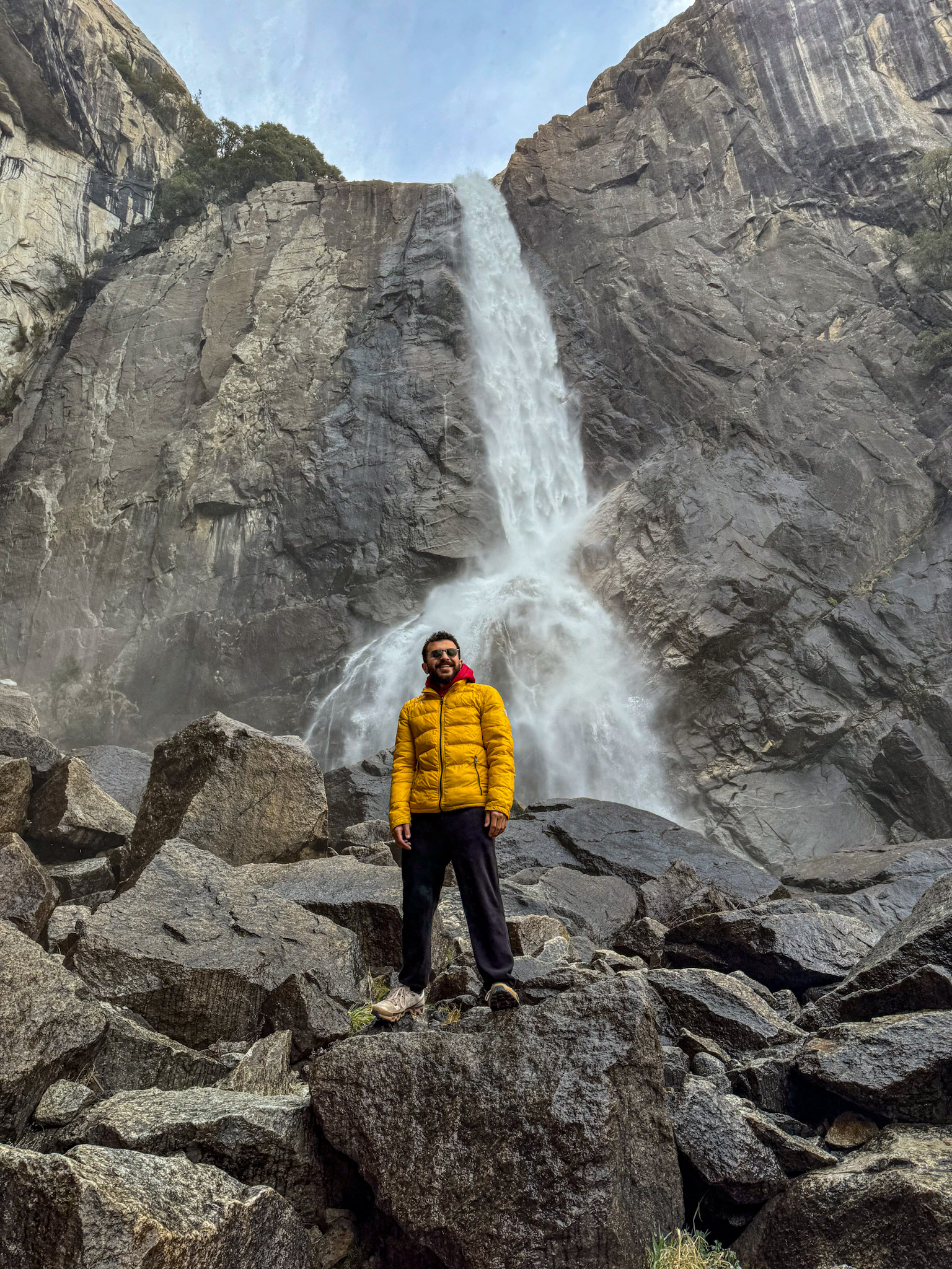 Person standing on rocks at the base of Yosemite Falls with waterfall crashing down granite cliff.