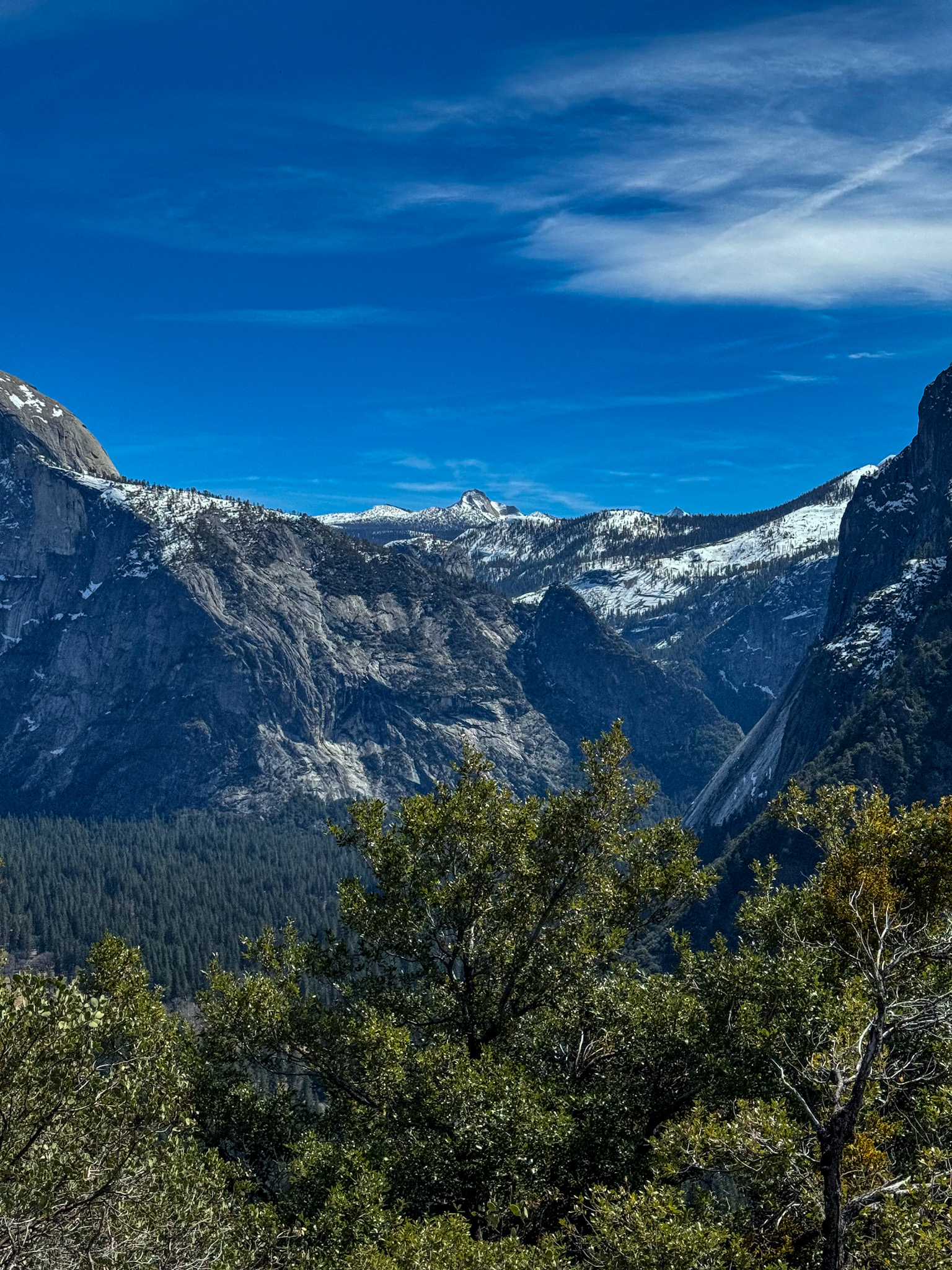 Yosemite National Park mountain landscape with snow-covered peaks, granite cliffs, and forest valley under blue sky.
