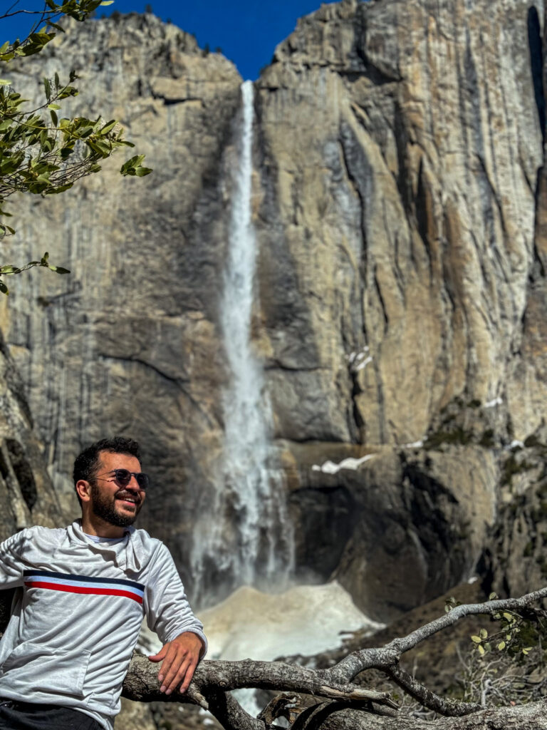 Person sitting near Yosemite Falls with tall waterfall cascading down granite cliff in Yosemite National Park.