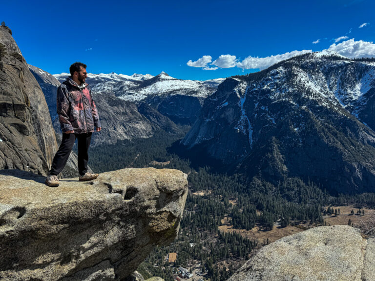 Person standing on cliff at Upper Yosemite Falls overlooking Yosemite Valley and snow-covered mountains.