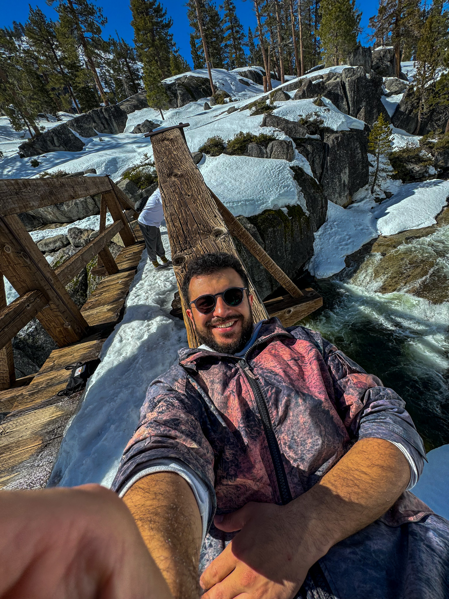 Person taking selfie on wooden bridge over flowing creek in snowy Yosemite mountain landscape.
