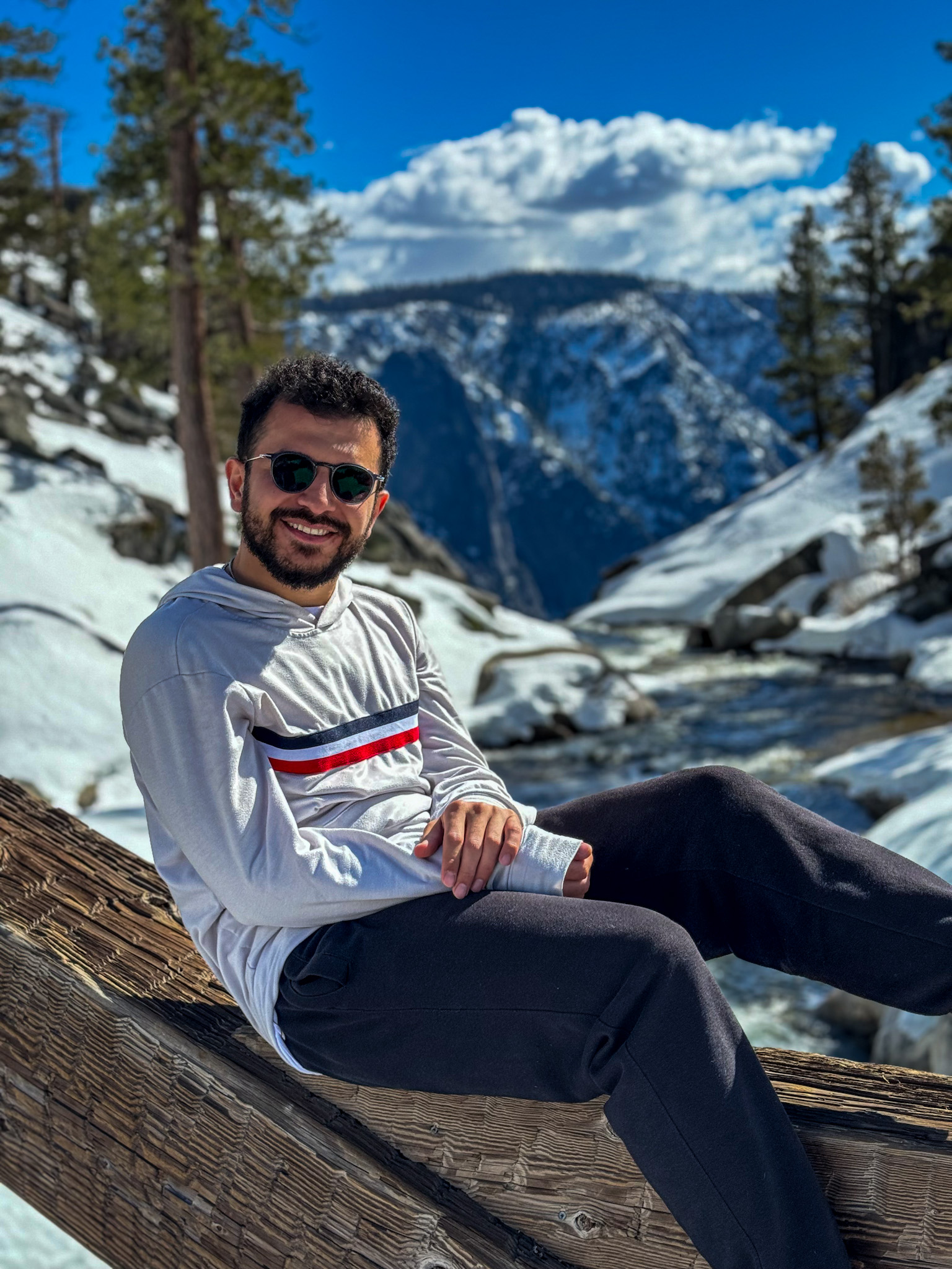 Person sitting by river in snowy Yosemite landscape with mountains and pine trees under blue sky.