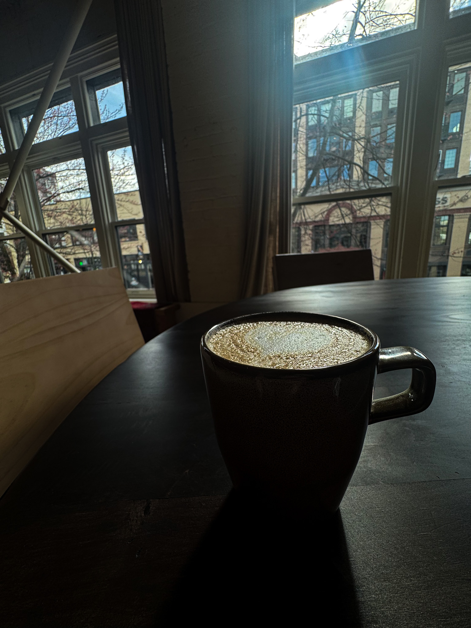 Cup of coffee on a dark table by a large window with city buildings visible outside.
