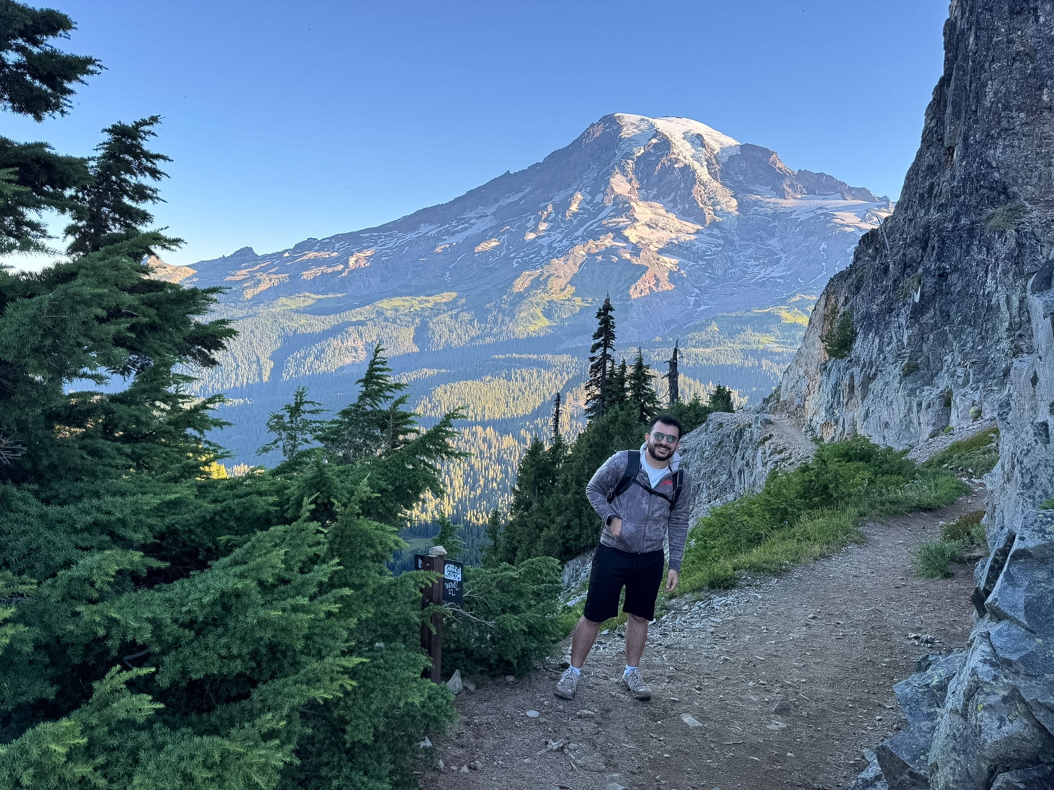 Hiker standing on Pinnacle Peak Trail with Mount Rainier in the background and rocky cliffs along the trail.