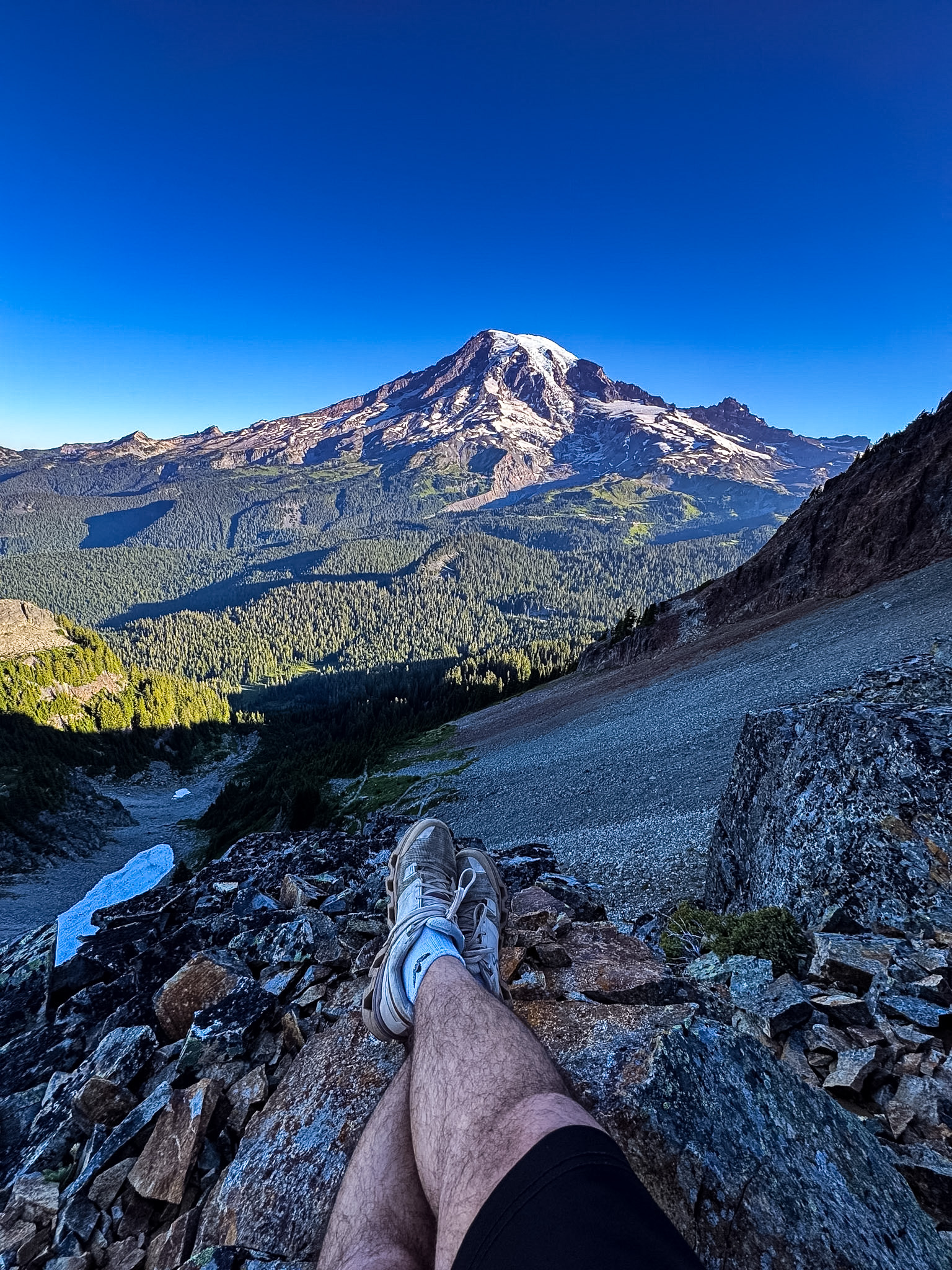 View of Mount Rainier from a rocky viewpoint on Pinnacle Peak Trail, with a hiker’s legs resting in the foreground.