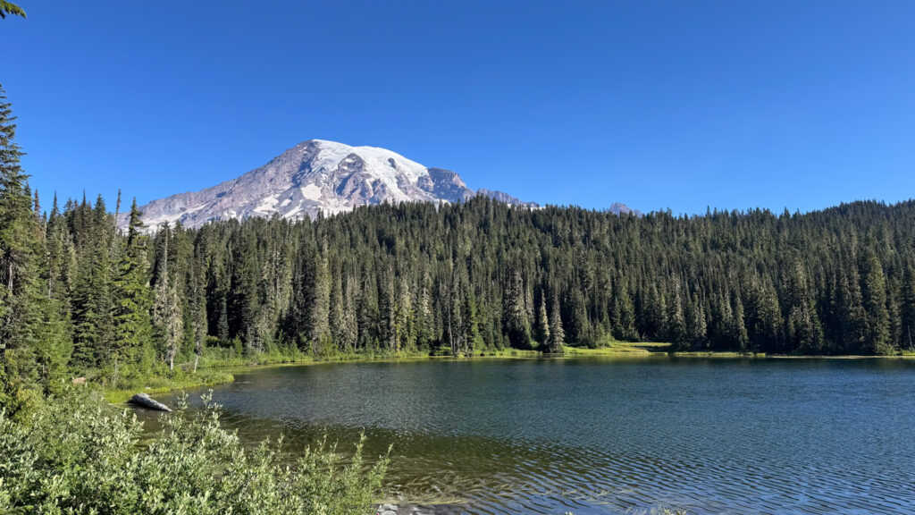 Alt text: Hiker standing on Pinnacle Peak Trail with Mount Rainier in the background and rocky cliffs along the trail.