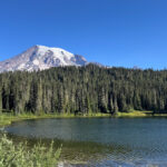 Alt text: Hiker standing on Pinnacle Peak Trail with Mount Rainier in the background and rocky cliffs along the trail.