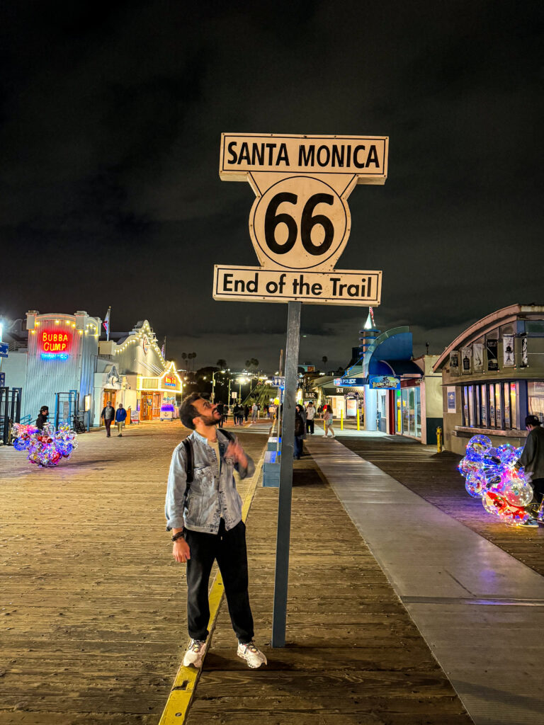 Traveler standing under the Route 66 End of the Trail sign on Santa Monica Pier at night.