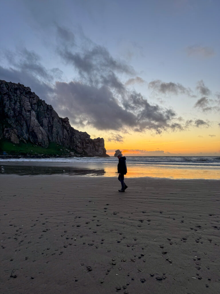 Person walking on the beach near Morro Rock during sunset in California.