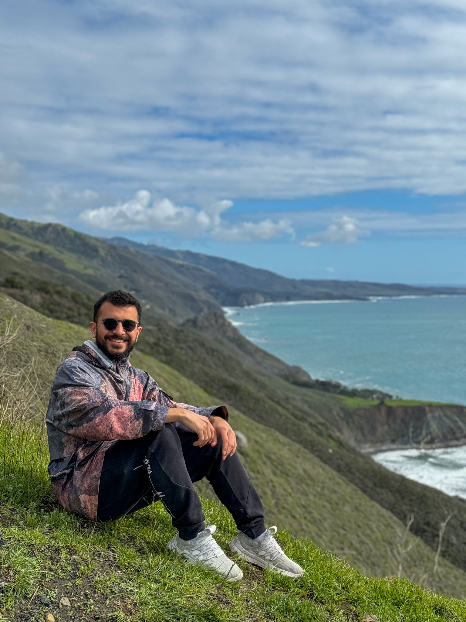 Traveler sitting on a grassy cliff overlooking the Big Sur coastline in California.