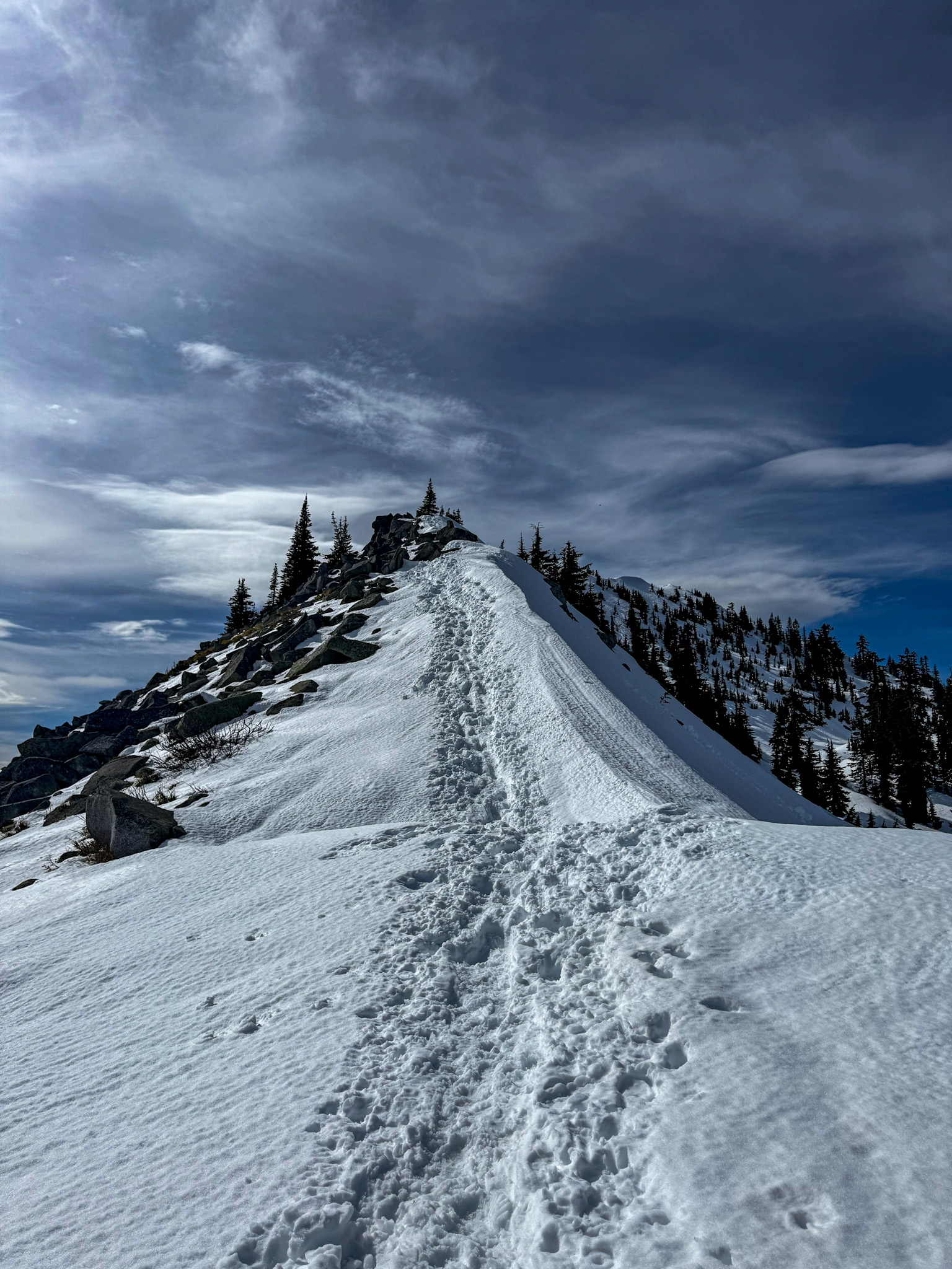 Snow-covered ridge trail near Granite Mountain summit with deep footprints and dramatic clouds in Washington.