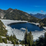 Partially frozen Snow Lake surrounded by evergreen forest and mountains in Snow Lake Hike Washington.
