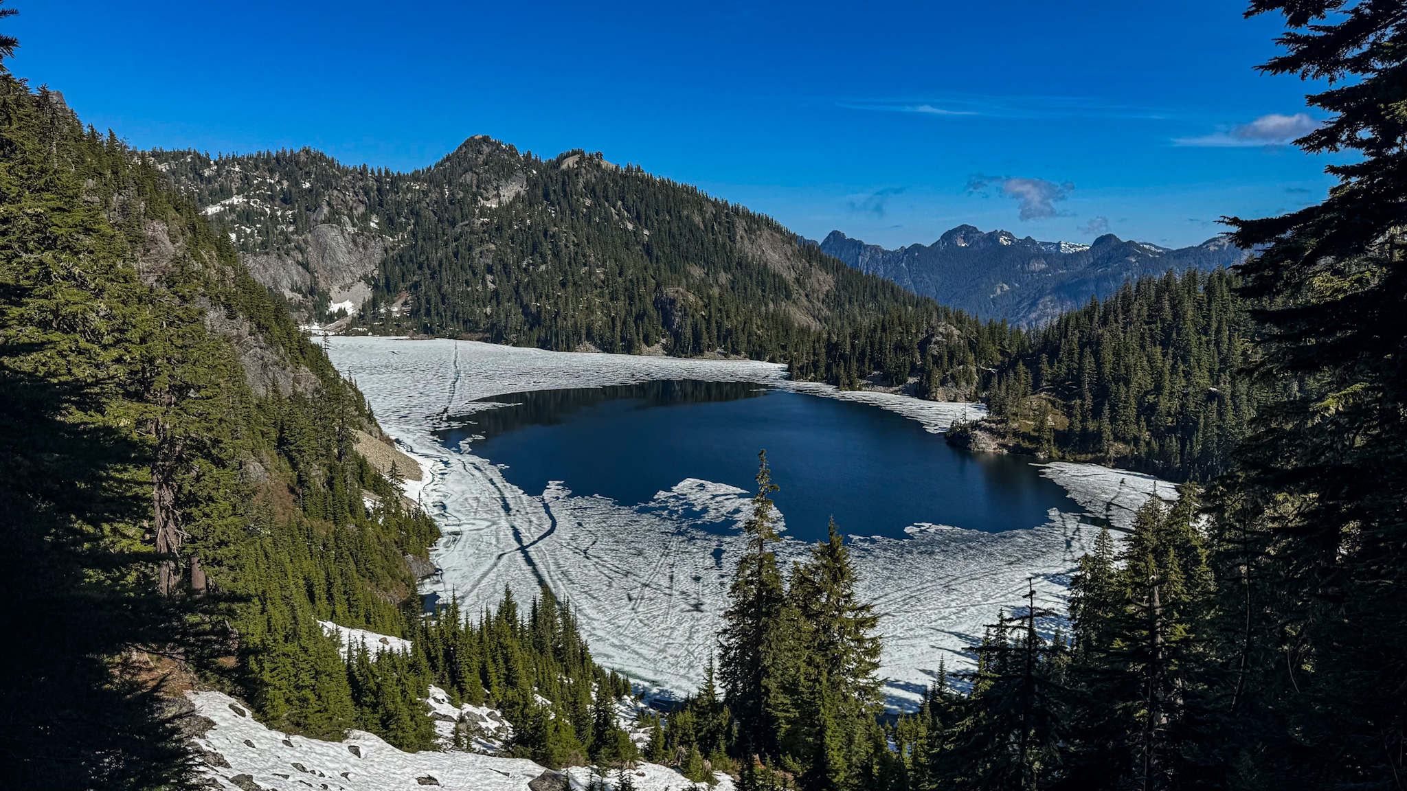 Partially frozen Snow Lake surrounded by evergreen forest and mountains in Snow Lake Hike Washington.