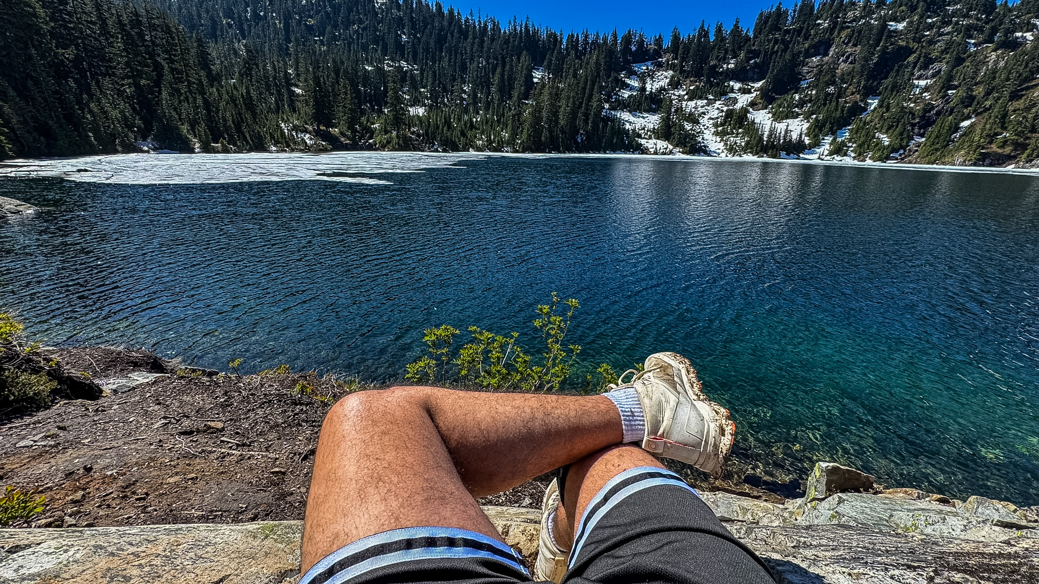 Hiker resting beside Snow Lake with clear blue water, lingering snow, and alpine forest near Snoqualmie Pass, Washington.